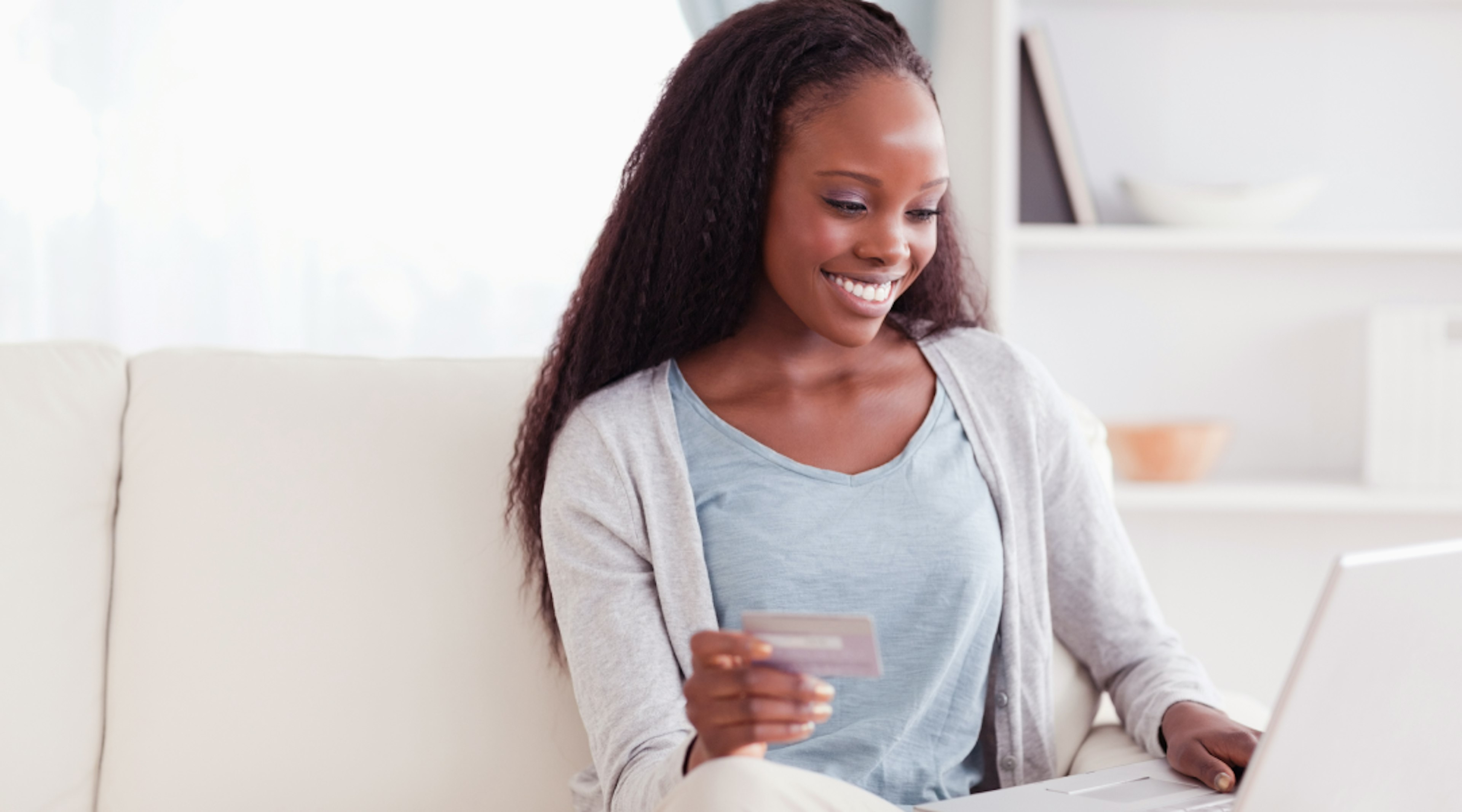 A woman sits on the couch holding her laptop in one hand and her credit card in the other.