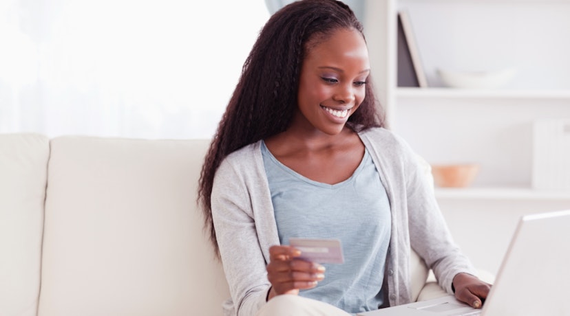 A woman sits on the couch holding her laptop in one hand and her credit card in the other.