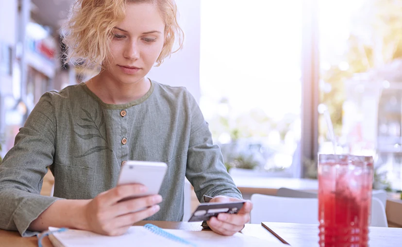 Woman looks down at her phone with a credit card in her other hand. She's sitting at a table with a dark red iced herbal blend.