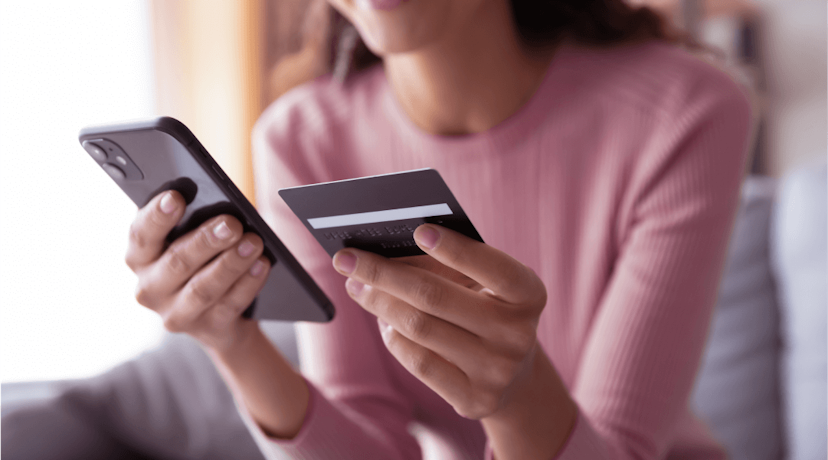 A woman sits on the couch holding her phone in one hand and her credit card in the other.