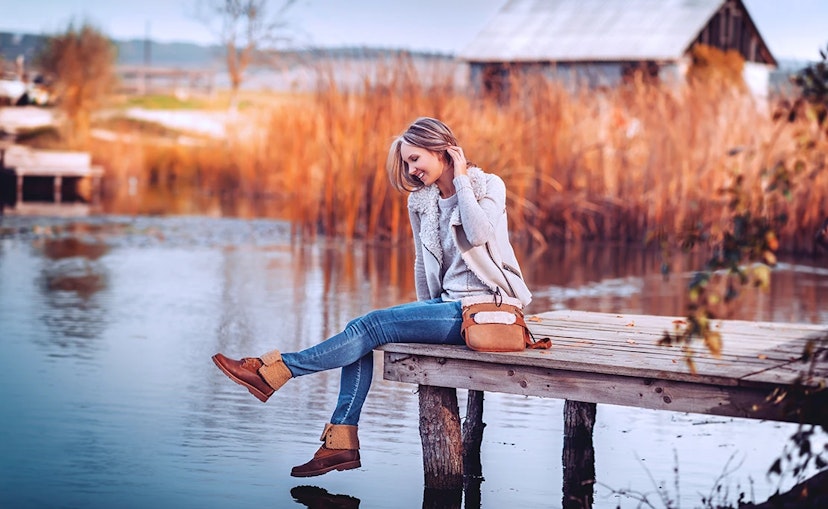 Woman sits on a pier over water. She wears jeans and a cream overcoat