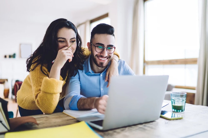 A couple sits at a table and looks at a laptop screen together