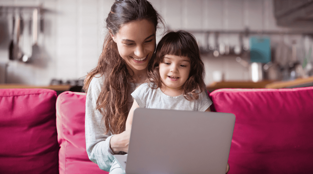 Woman on couch sits with child in lap. They're looking at a laptop on a coffee table before them.